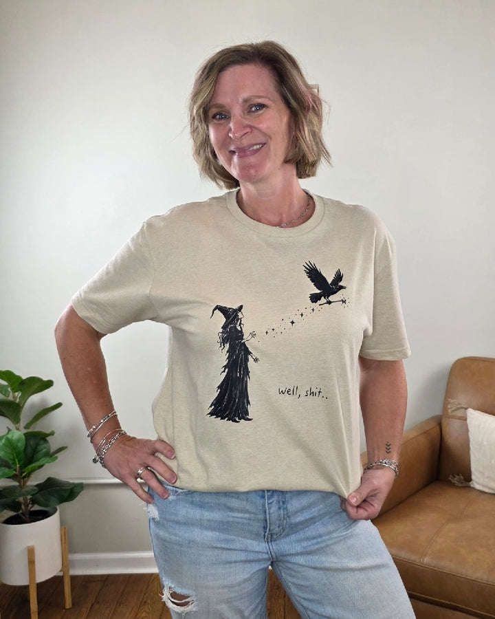 A woman wearing a tan Cosmos + Clover Boutique Witch Graphic Tee featuring a witch and a flying crow graphic with the text well, shit.. printed next to them.