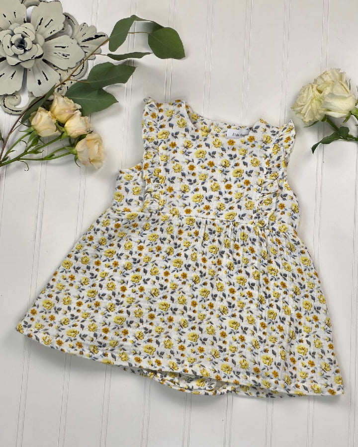 The Angel Dear Yellow Country Roses - Ruffle Bubble Dress, a sleeveless white baby dress in organic cotton muslin with a yellow floral pattern, is displayed flat on a white wooden surface with white roses and green leaves.