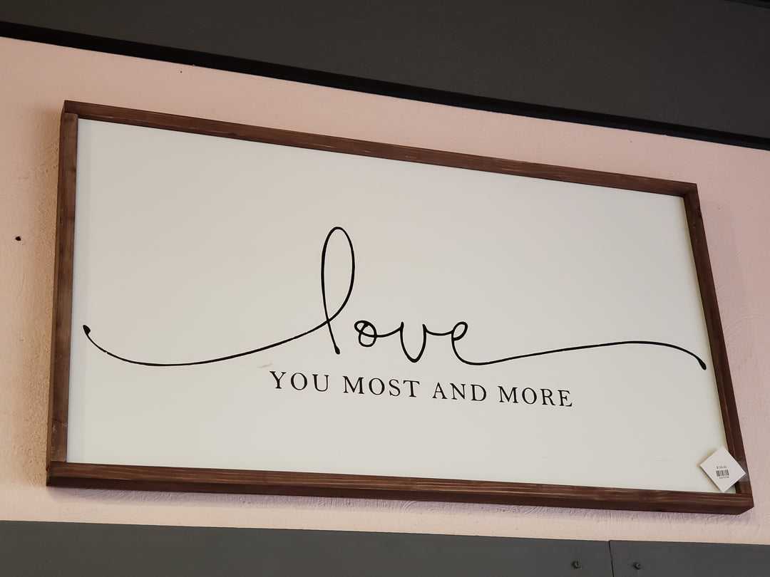 The “Love” Wood Sign by Cosmos and Clover features “Love you most and more” in black on white with a wooden frame, displayed on a light pink wall. Ideal for local pick-up.