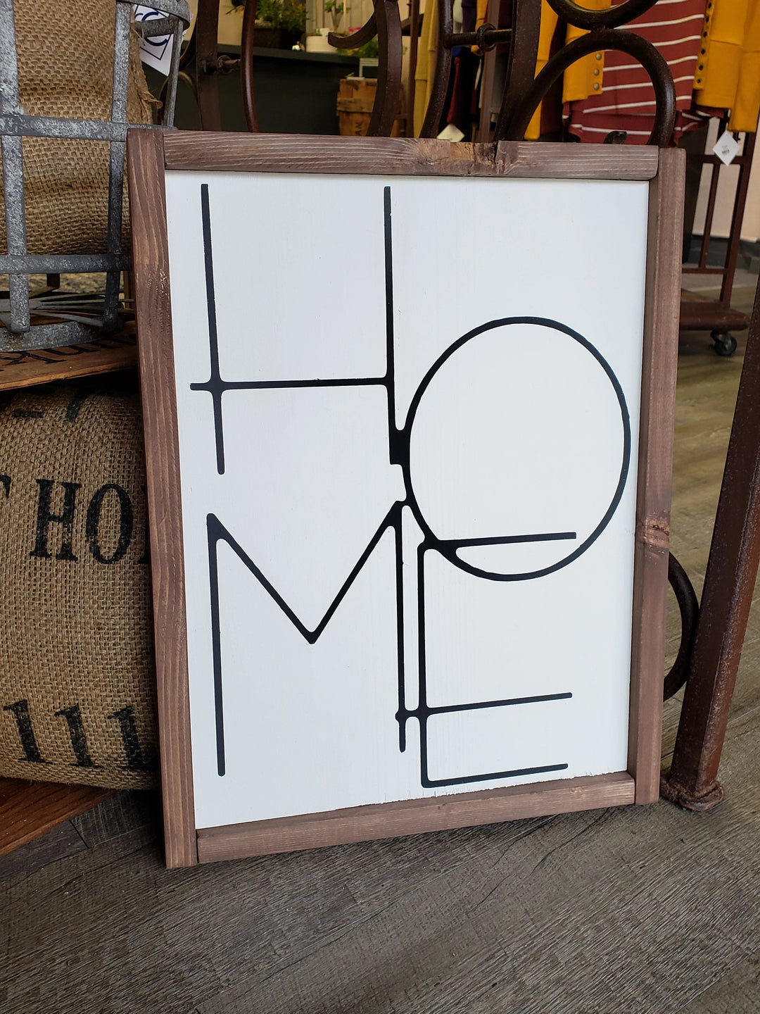 The "HOME" Wood Sign by Cosmos and Clover features a modern minimalist black font, with the O overlapping H and E. Framed and painted wood, it rests against a rustic in-store display. Local pick-up available.
