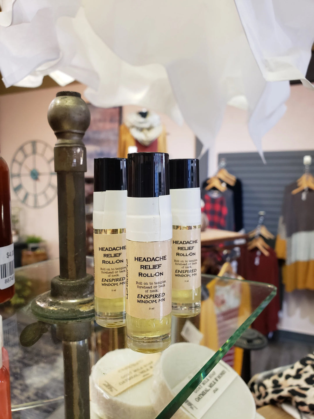 Three bottles of Enspired Headache Relief Roll-on are displayed on a glass shelf in a boutique, with blurred clothing racks and a decorative lamp in the background.