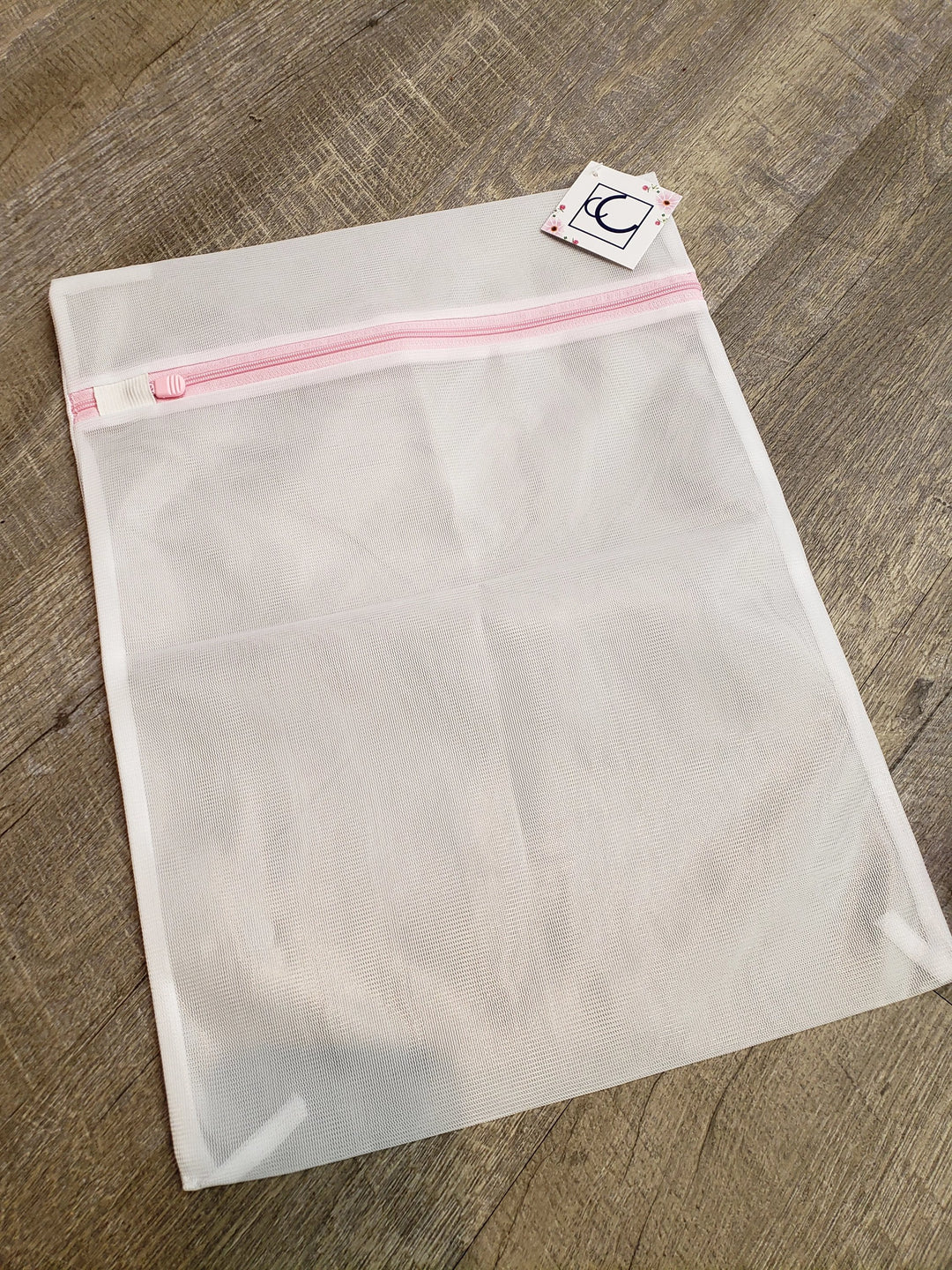 The Amazon Medium Mesh Laundry Bag, measuring 15.5x11.5 inches, features a pink zipper and a small logo tag attached, shown lying flat on a wooden floor.