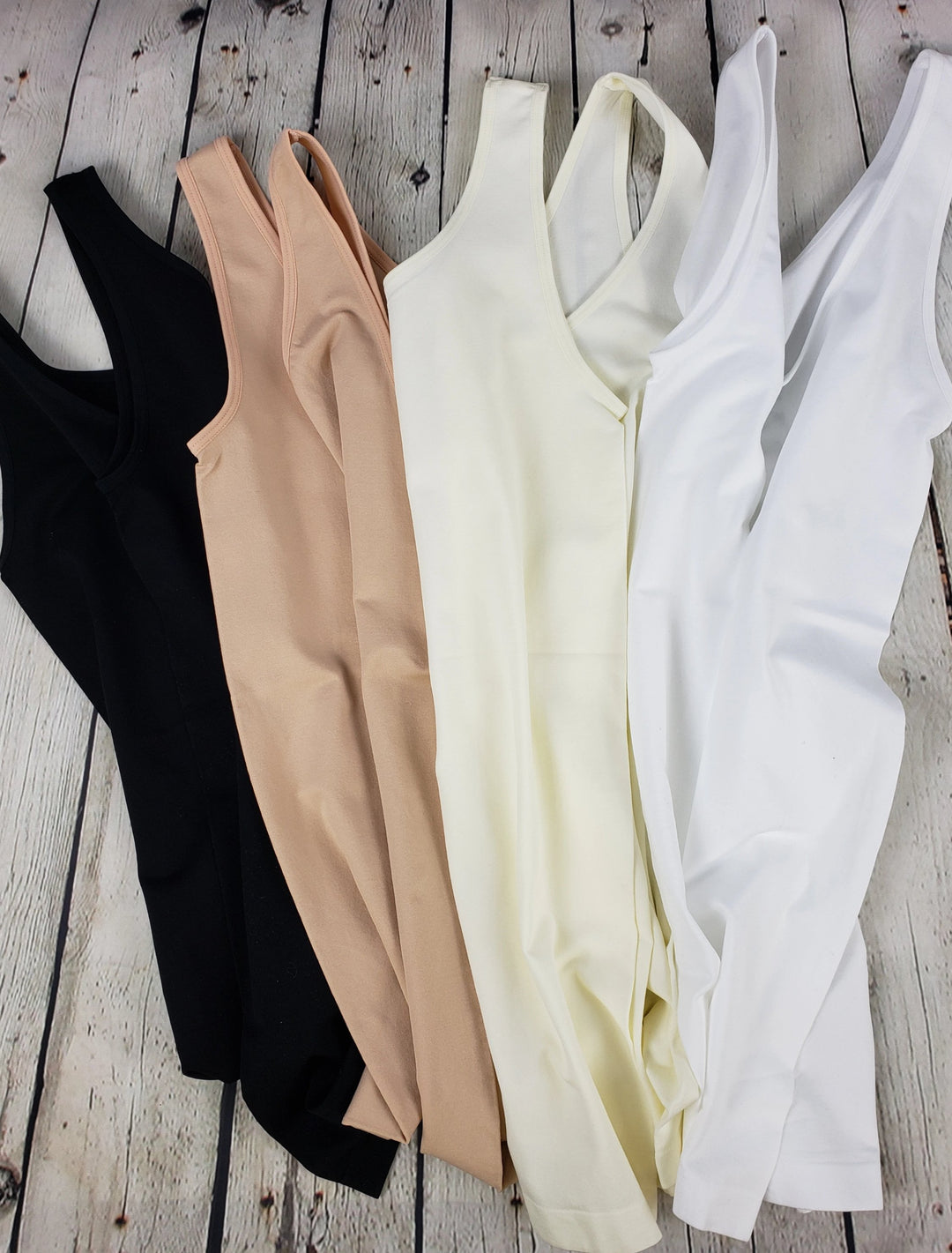 Five Yelete Group Wide Strap Layering Tanks in black, beige, light beige, ivory, and white are arranged side by side on a light wooden surface—ideal seamless basics for layering any outfit.