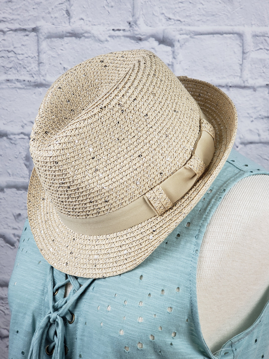 The Too Too Hat Straw Hat in beige natural fiber, featuring a matching ribbon band, is displayed on a mannequin wearing a light blue sleeveless lace-up top against a white brick background.