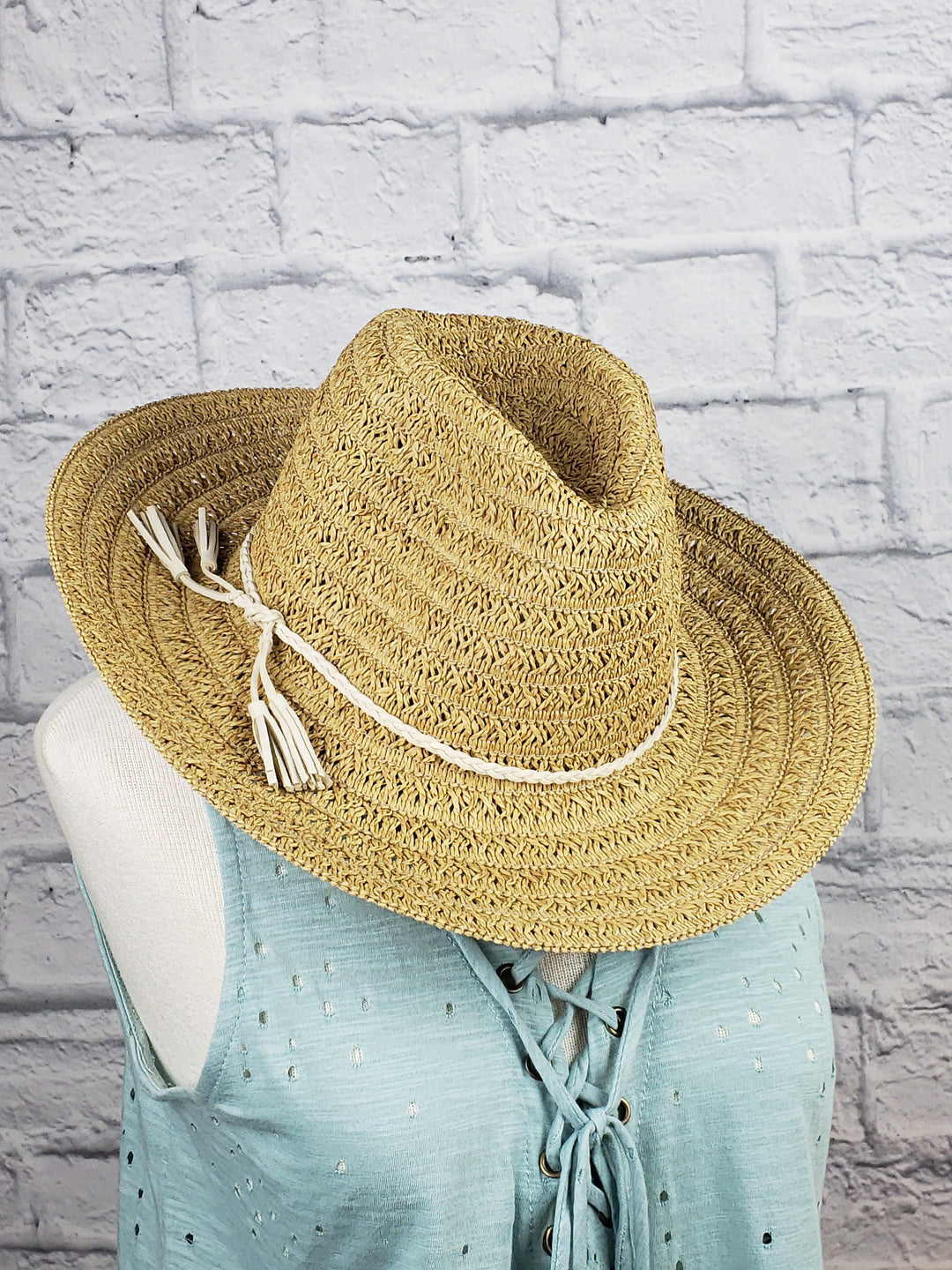 A brown Too Too Hat Panama Hat with a white cord and tassels is displayed on a mannequin in a light blue sleeveless lace-up top, set against a white brick wall.