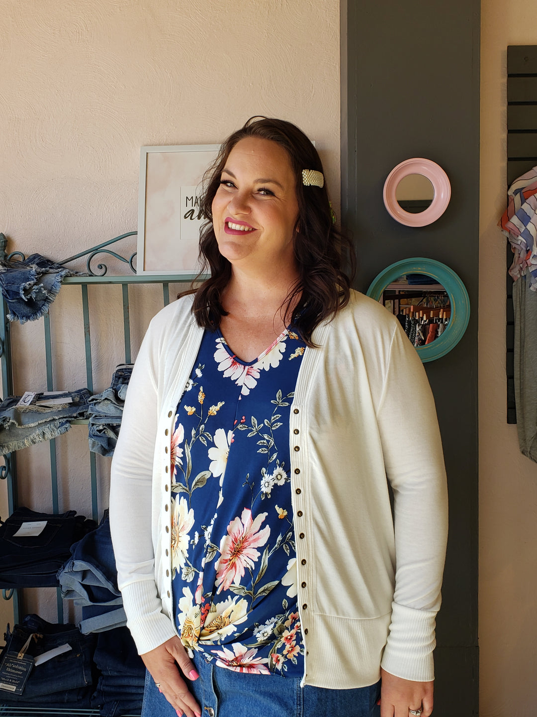 A woman with wavy brown hair smiles indoors, wearing the Zenana Long Sleeve Snap Front Cardi - Extended Sizes over a blue floral top and jeans. Shelves with folded denim and a decorative mirror are visible behind her.