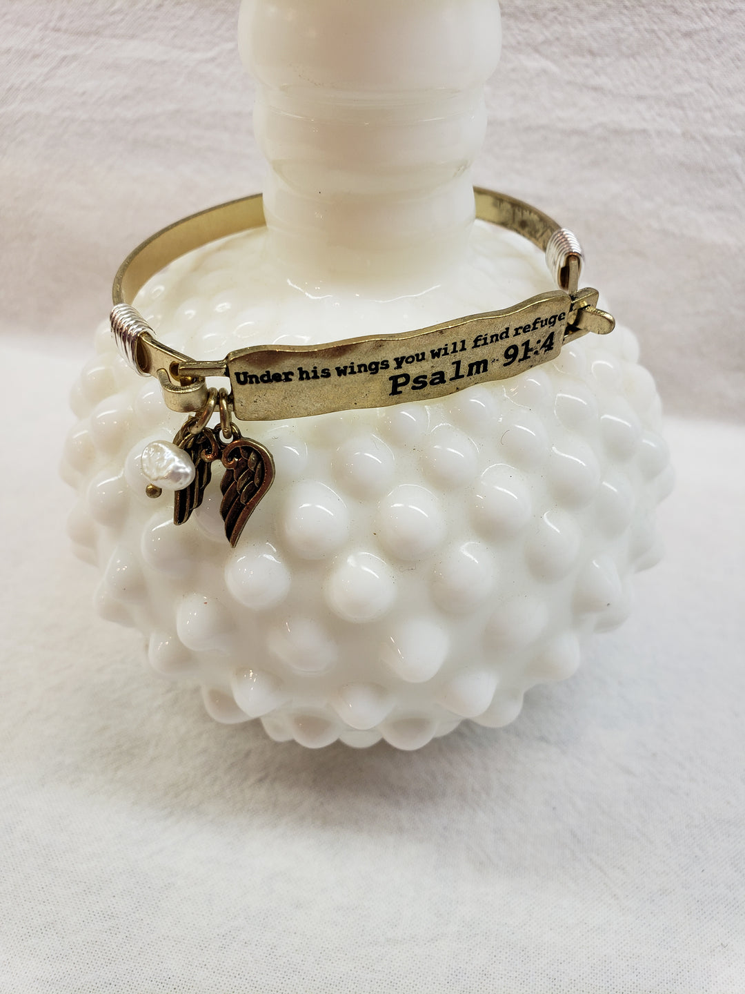 The Cash + Carry Metal Bangles feature a gold finish, a Psalm 91:4 engraved plate, wing and pearl charms, and rest on a white textured vase. Pair with other bangles for assorted colors.