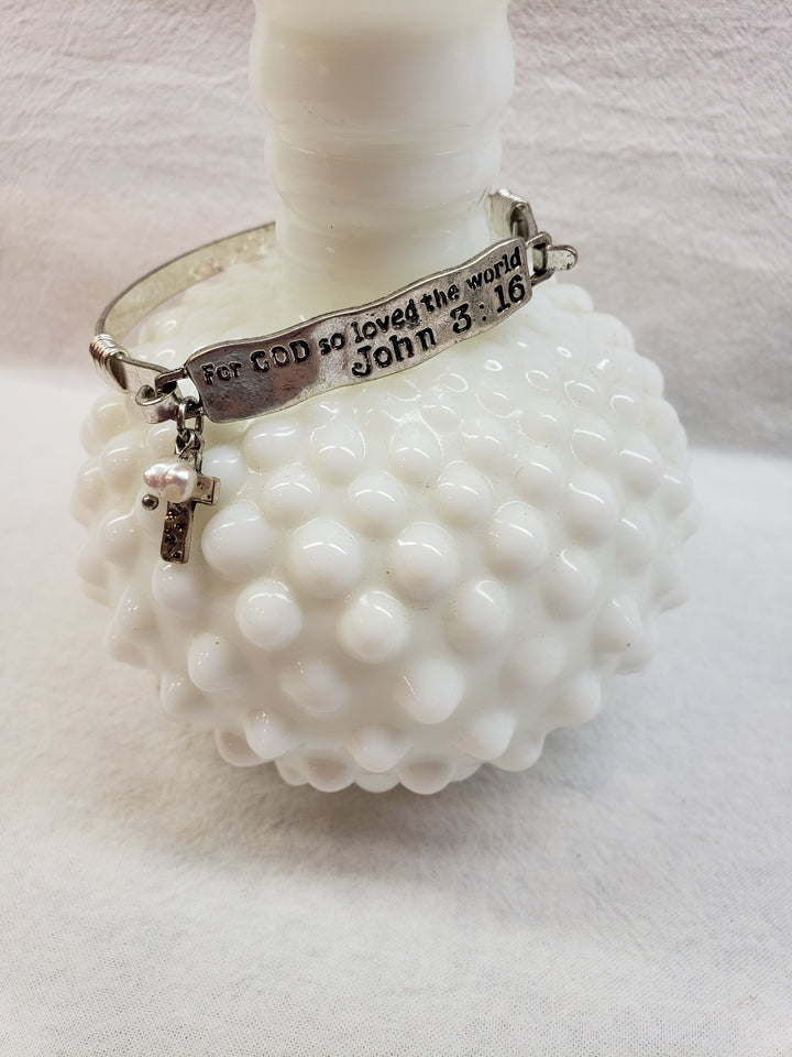 The Cash + Carry Metal Bangles feature a silver bangle with a cross charm and "For GOD so loved the world John 3:16" engraved. Shown on a white glass vase, they're perfect for stacking with other colorful metal bangles.