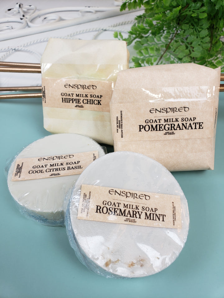 Four bars of Enspired Handmade Soap in scents like Hippie Chick, Pomegranate, Cool Citrus Basil, and Rosemary Mint are wrapped and labeled, displayed on a light blue surface with a green plant in the background.