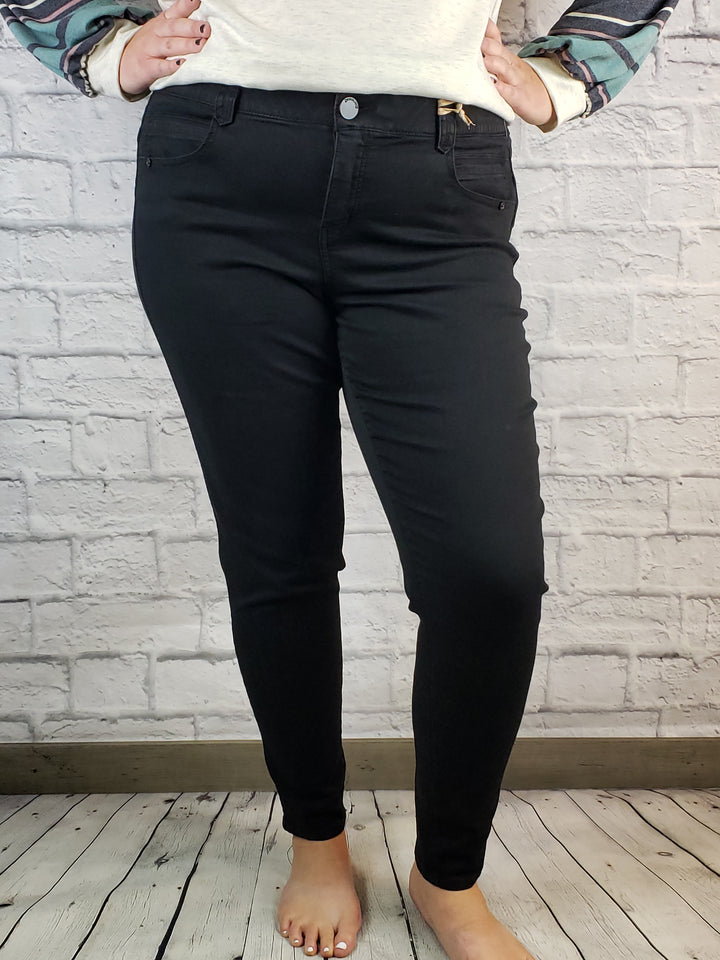 A person wearing Democracy "Ab"solution Skinny: Black - Ext. Sizes jeans stands barefoot on a wooden floor against a white brick wall, hands on hips, paired with a cream sweater.