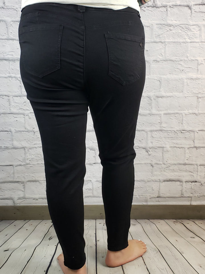 A person stands barefoot on a wooden floor, facing a white brick wall and wearing Democracy’s "Ab"solution Skinny: Black - Ext. Sizes jeans with back pockets and seams, paired with a white top. The image is taken from behind to show the fit.