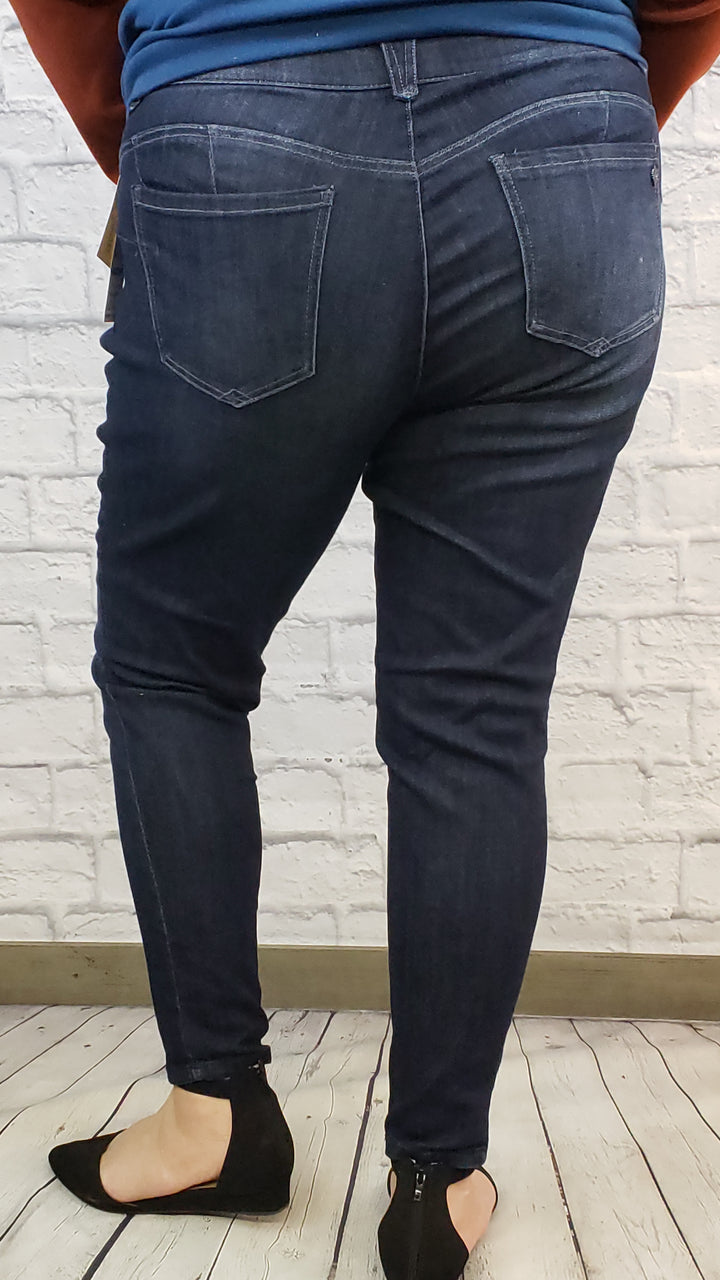 A person stands with their back to the camera, wearing Democracy "Ab"solution Skinny Jeans with Distressing: Extended Sizes and black flats, on a light wooden floor in front of a white brick wall.