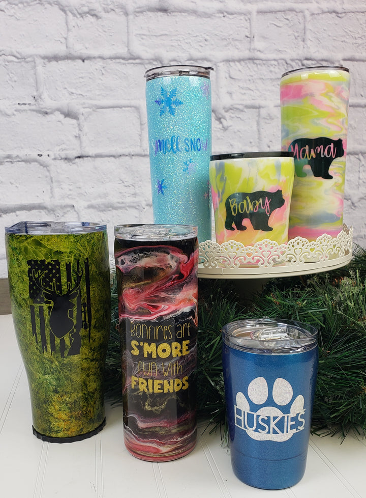 A set of six Decorated Mugs by Adam & Perian Crafts, each with unique designs like animals, text, and marble patterns, displayed on a wooden surface with greenery and a white brick wall backdrop.