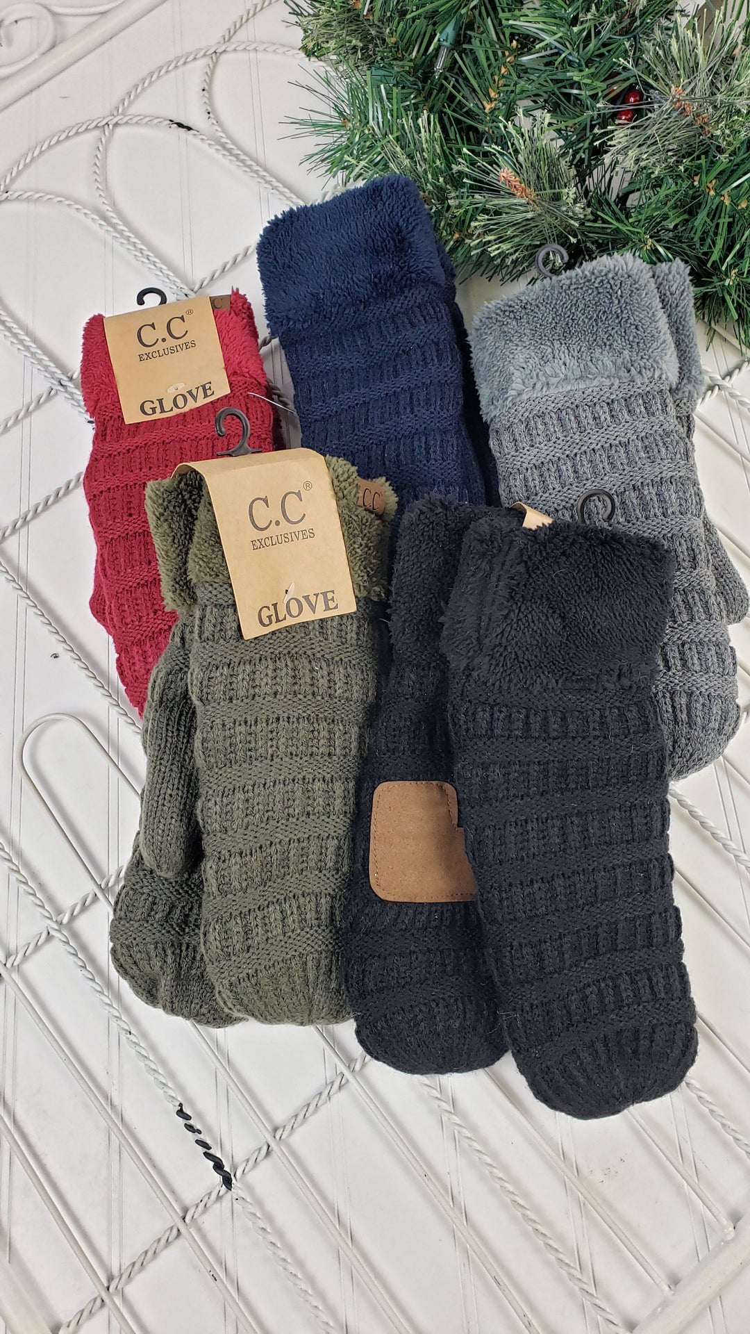 Five pairs of CC Beanie Mittens ($15 each) from CC Beanie in red, navy, gray, olive green, and black are arranged on a white surface beside a green holiday wreath. Each mitten features the CC Beanie brand label.