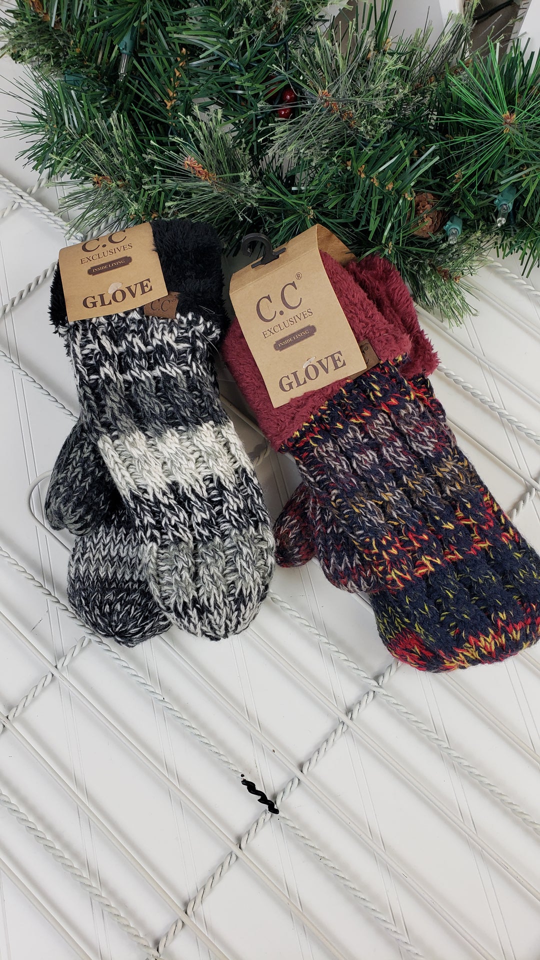 Two pairs of CC Beanie Mittens ($18) with brown labels rest on a white surface under a green holiday garland—one black and white, one maroon and multicolored—both from CC Beanie’s Winter Accessories collection.