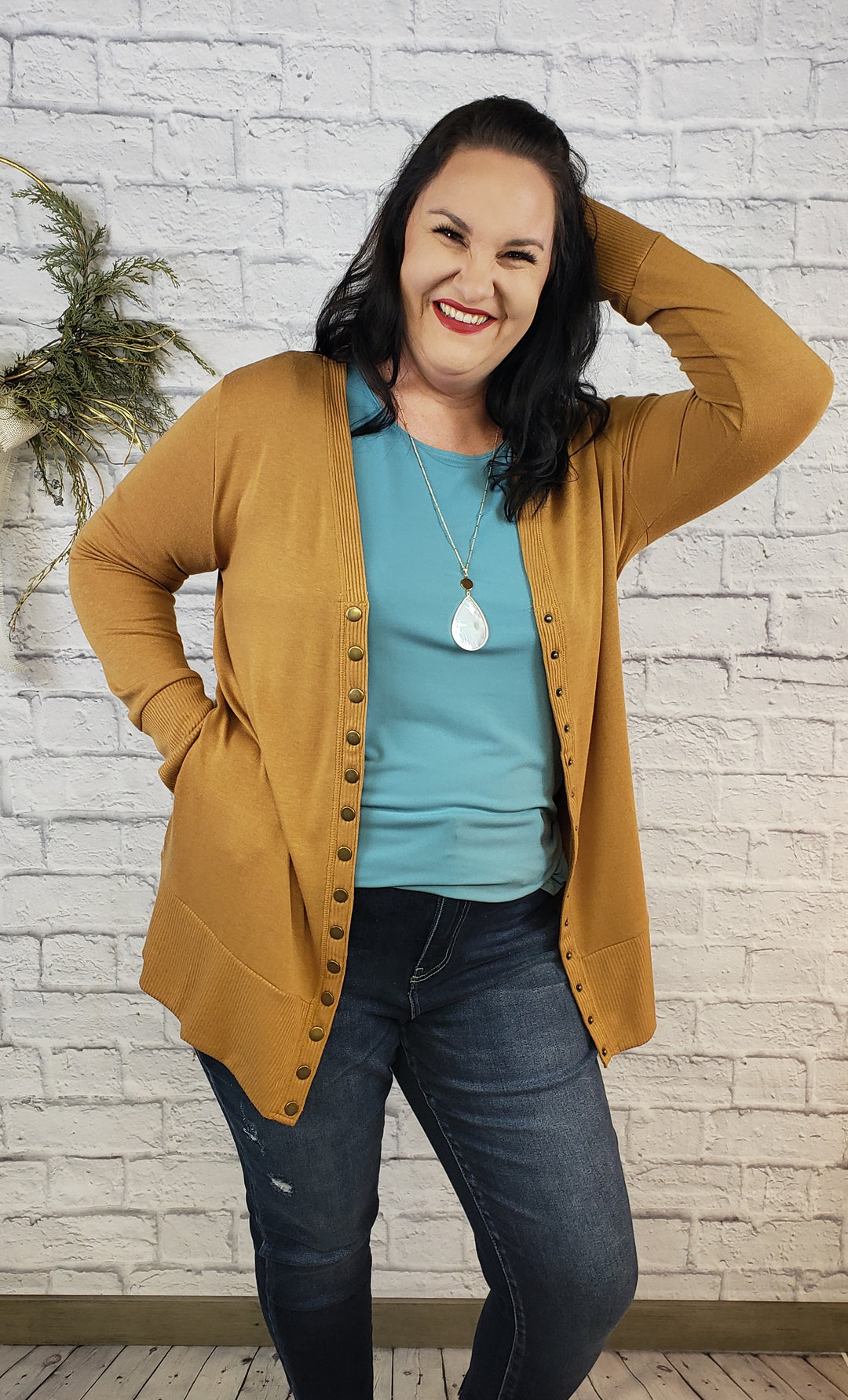 A woman with long dark hair smiles while posing in front of a white brick wall, wearing the Zanana Long Sleeve Snap Cardi with Pockets over a teal shirt and jeans. She stands beside a small plant decoration, accessorized with a pendant necklace.