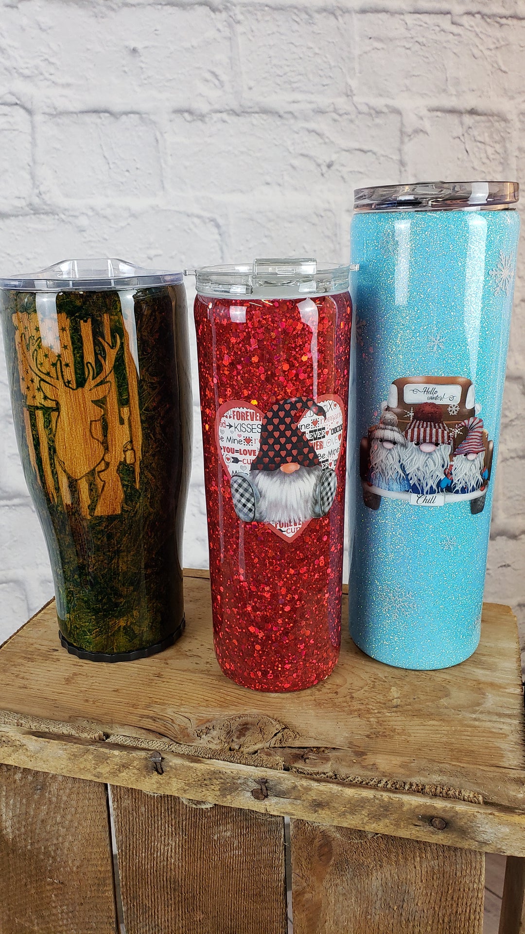 Three Decorated Mugs by Adam & Perian Crafts—featuring a forest print, a red glittery gnome with hearts, and a light blue holiday cake—are displayed on wood against a white brick wall, perfect for mug enthusiasts.