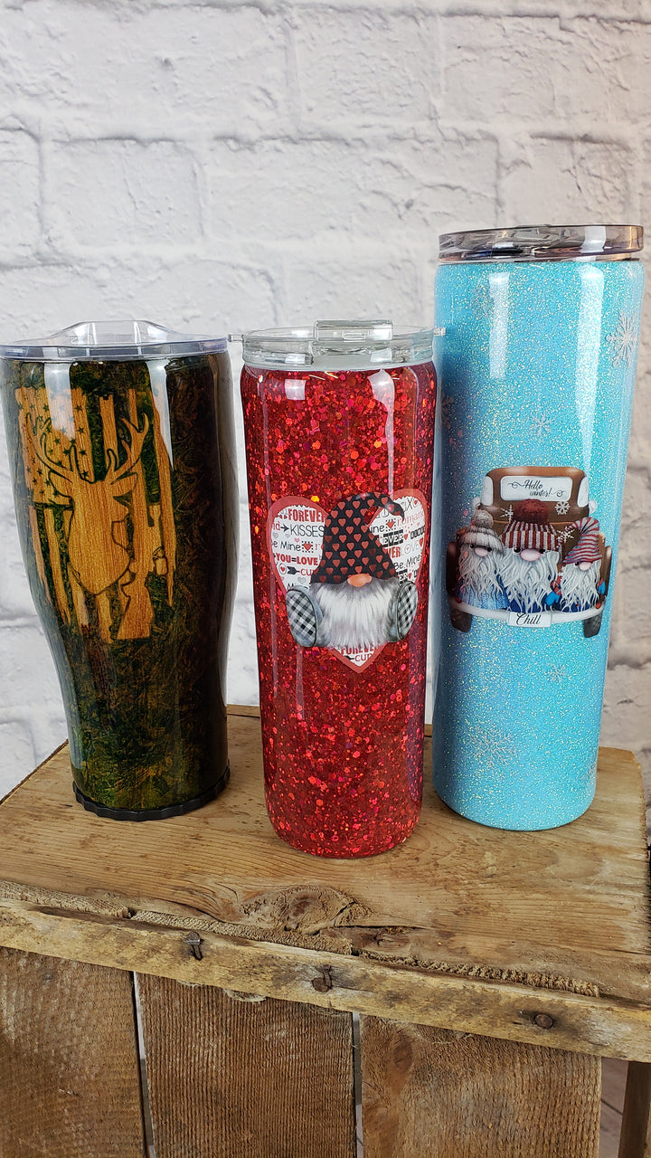 Three Decorated Mugs by Adam & Perian Crafts—featuring a forest print, a red glittery gnome with hearts, and a light blue holiday cake—are displayed on wood against a white brick wall, perfect for mug enthusiasts.