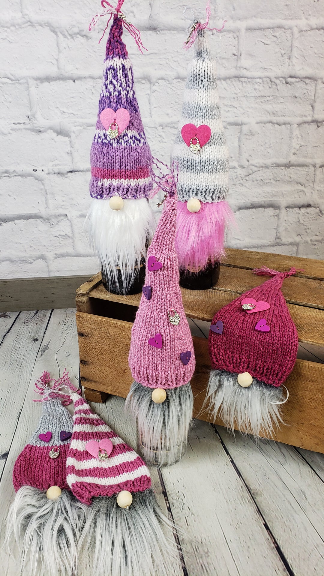 Five Nanny K's Kitchen Kreations Gnome Bottle Covers, each with a long white beard, round nose, and assorted pink, white, or purple knitted hats, are displayed on and in front of a wooden crate against a white brick wall.