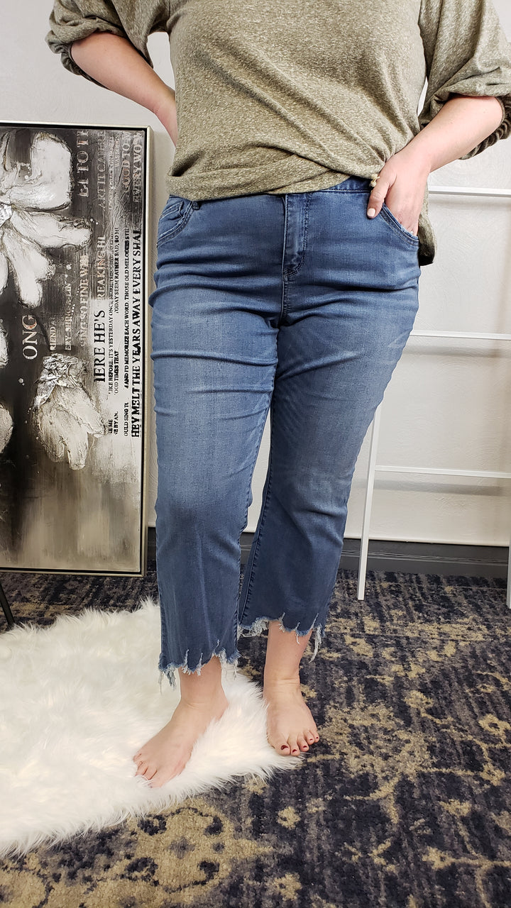 A person stands barefoot on a white rug, wearing blue Democracy "Ab"Solution Chewed Hem Crops (Extended Sizes) and a greenish-gray long-sleeve shirt, with one hand in their pocket. A decorative canvas leans against the wall nearby.