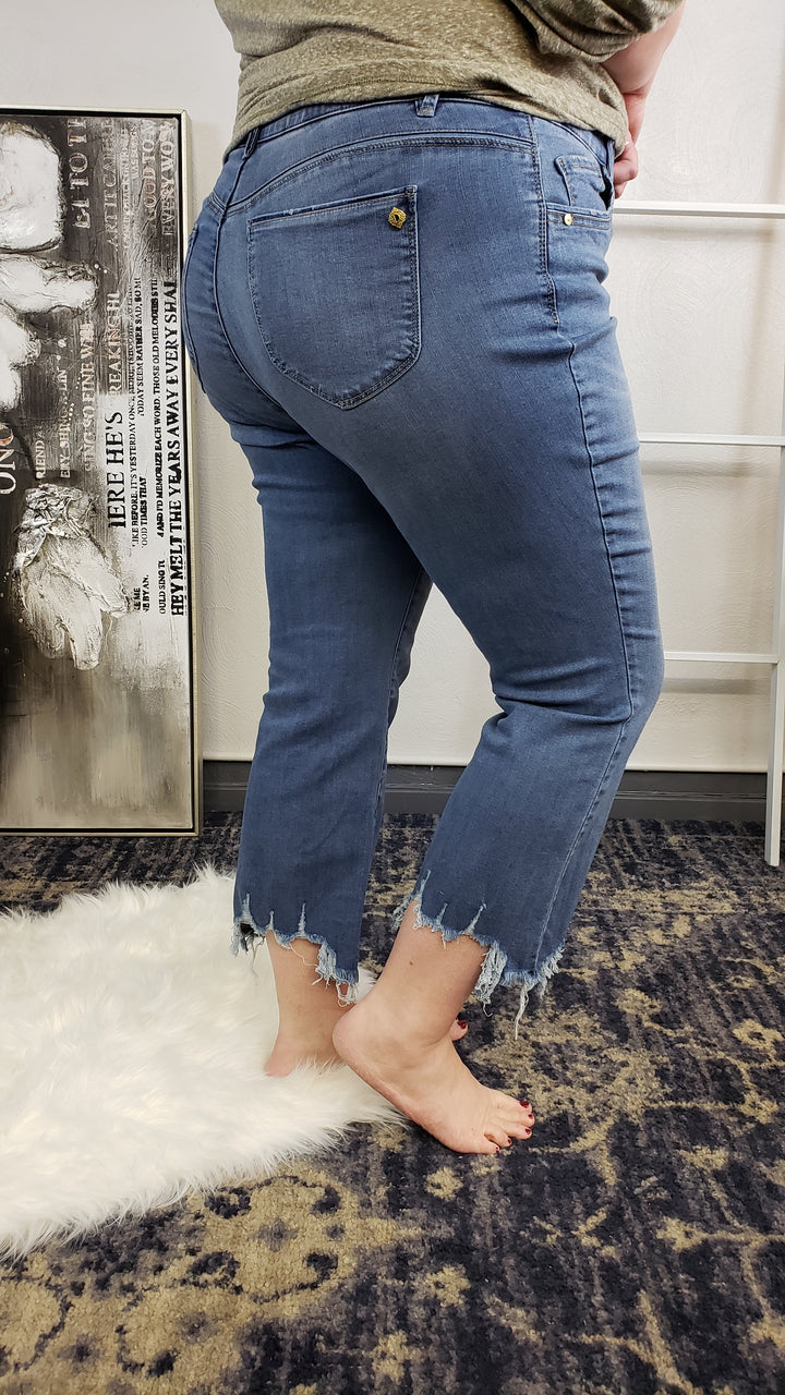 A person stands barefoot on a rug, wearing Democracy "Ab"Solution Chewed Hem Crops - Extended Sizes and a green top. The photo is taken from behind, focusing on the lower body, with a painting resting against the wall in the background.