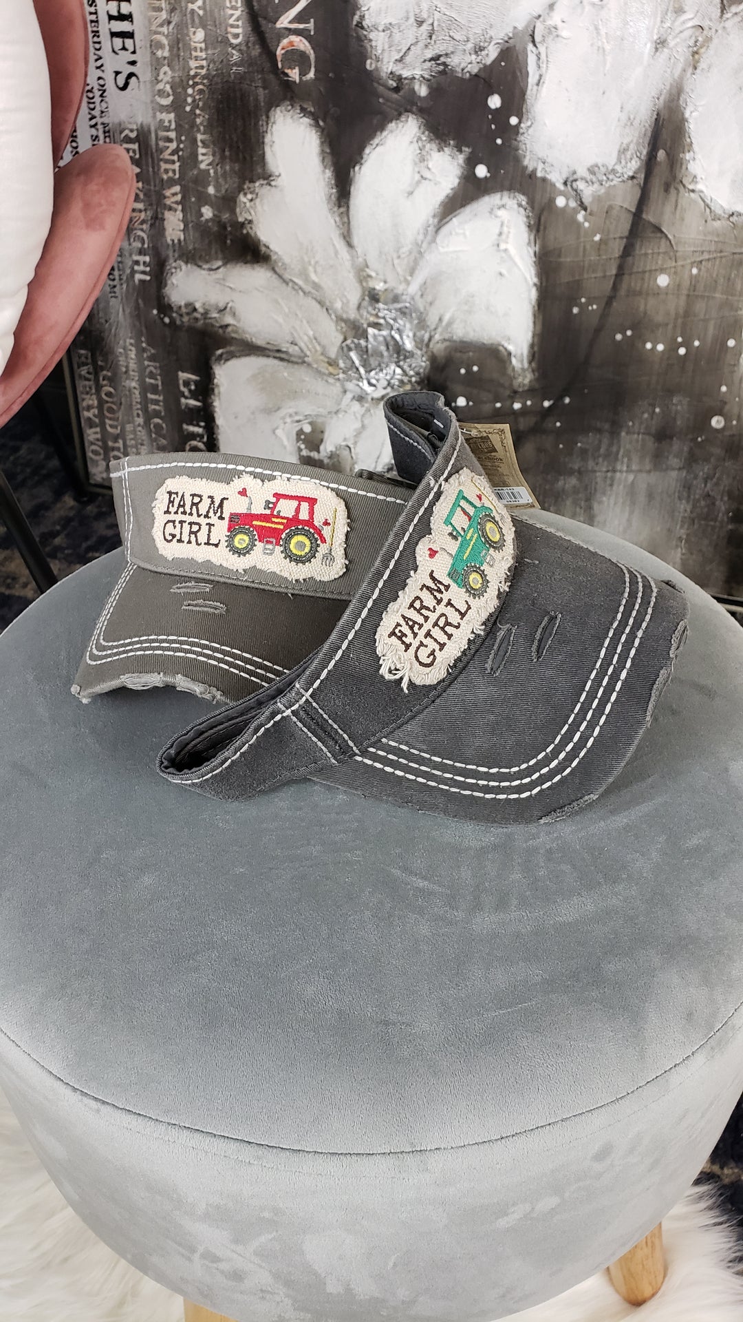 Two Hana Distressed Visors, each with a FARM GIRL patch—one red tractor, one green—are displayed on a soft gray ottoman. An assorted floral painting decorates the background.