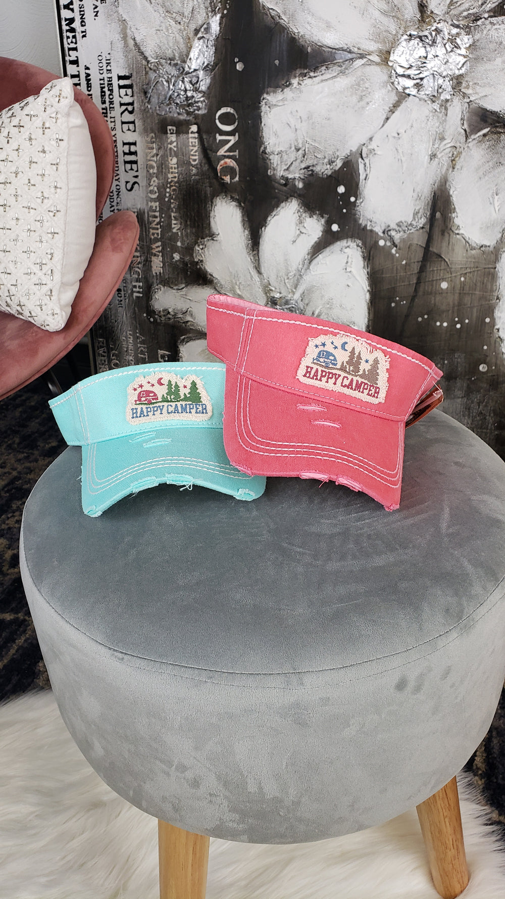 Two Hana Distressed Visors, one teal and one pink with "Happy Camper" text and camping graphics, sit on a round gray stool. In the background, a hand holds a small patterned pillow while abstract art decorates the wall.
