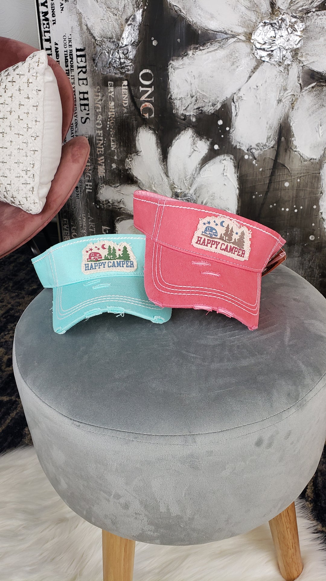 Two Hana Distressed Visors, one teal and one pink with "Happy Camper" text and camping graphics, sit on a round gray stool. In the background, a hand holds a small patterned pillow while abstract art decorates the wall.