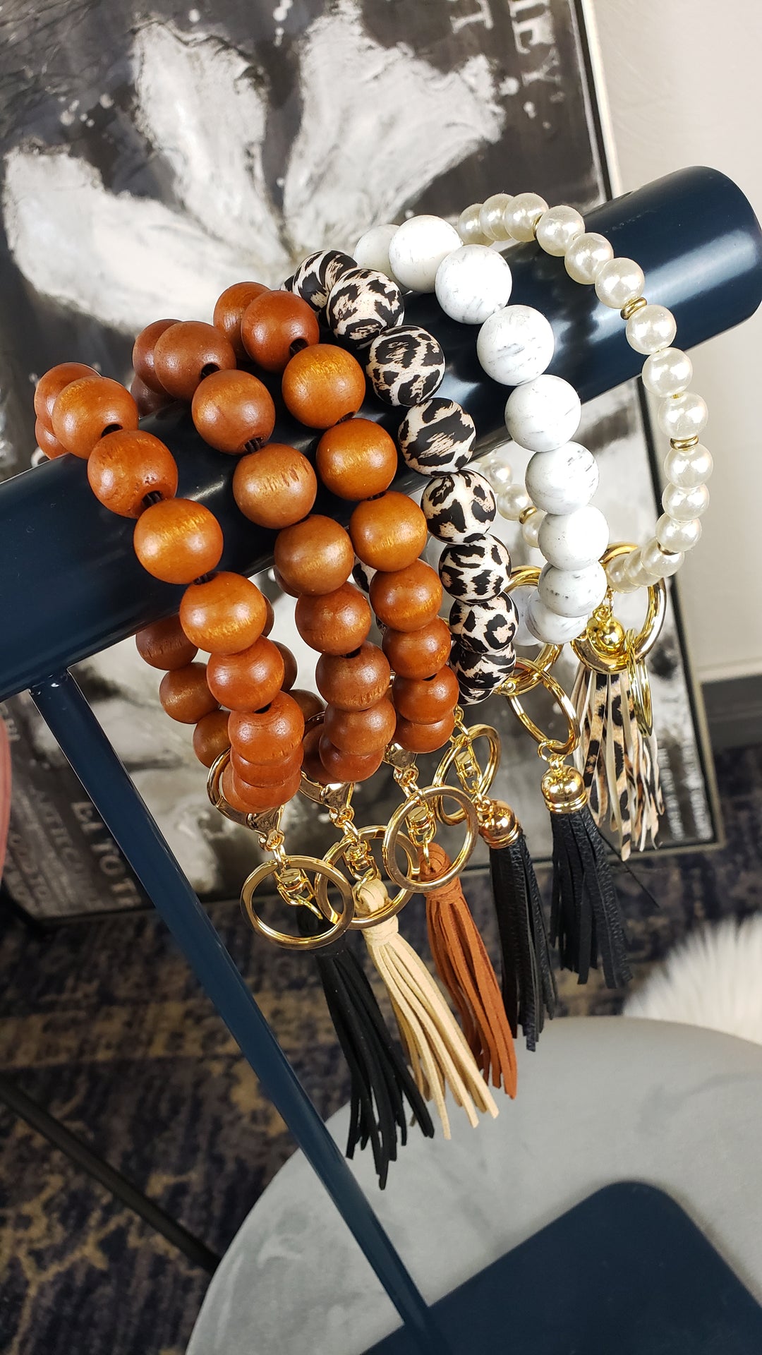 Four Aili's Corner Beaded Key Ring Bracelets—wooden, leopard print, white marble, and pearl-like—hang on a rod. Each bracelet has gold clasps plus either a tassel or a patterned charm at the end.