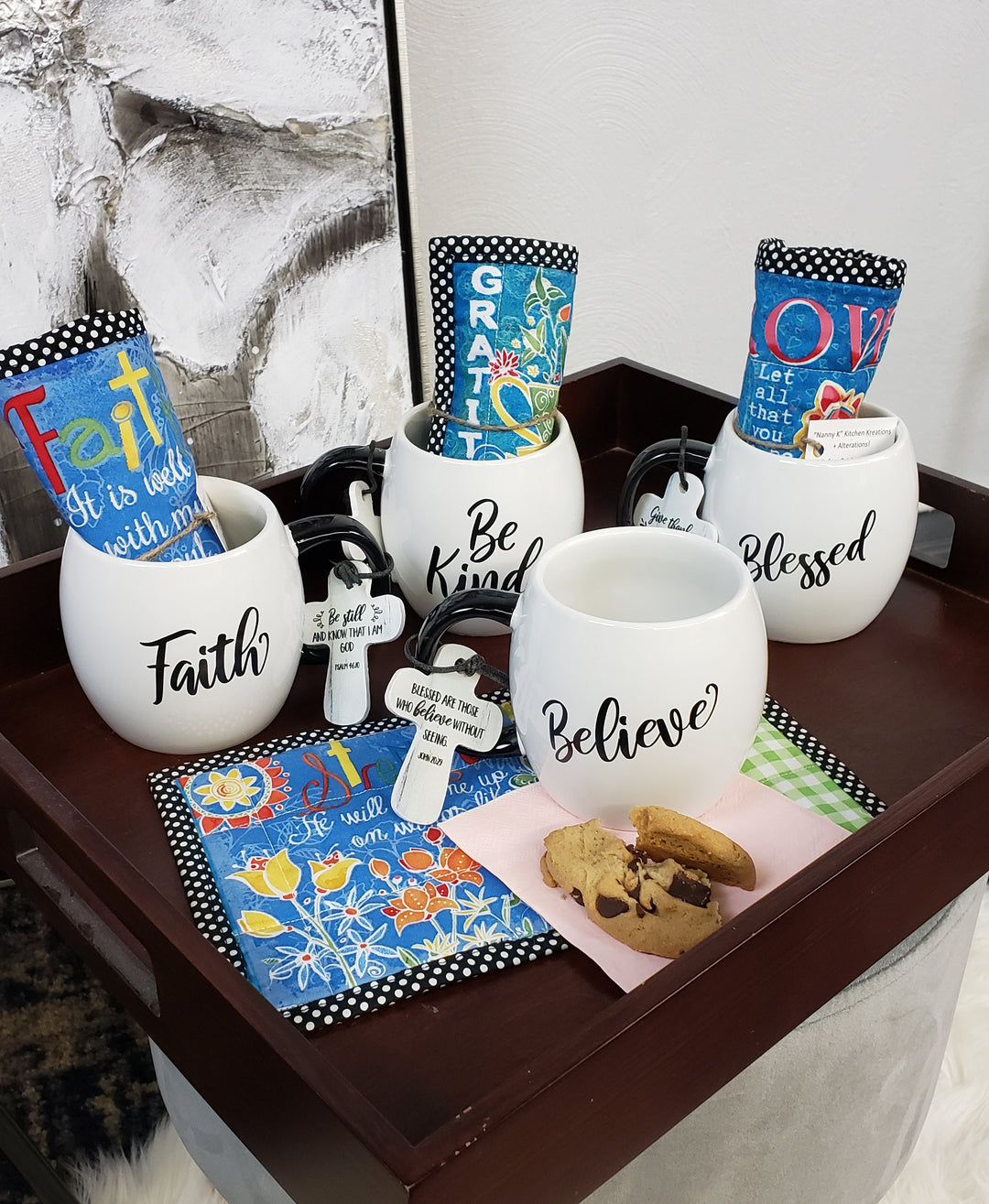 Nanny K's Kitchen Kreations Mug & Rug Sets feature a wooden tray with handmade mugs labeled Faith, Be Kind, Believe, Blessed, and Gratitude. Each mug includes colorful cloths with positive words and cookies on pink and green napkin "rugs.