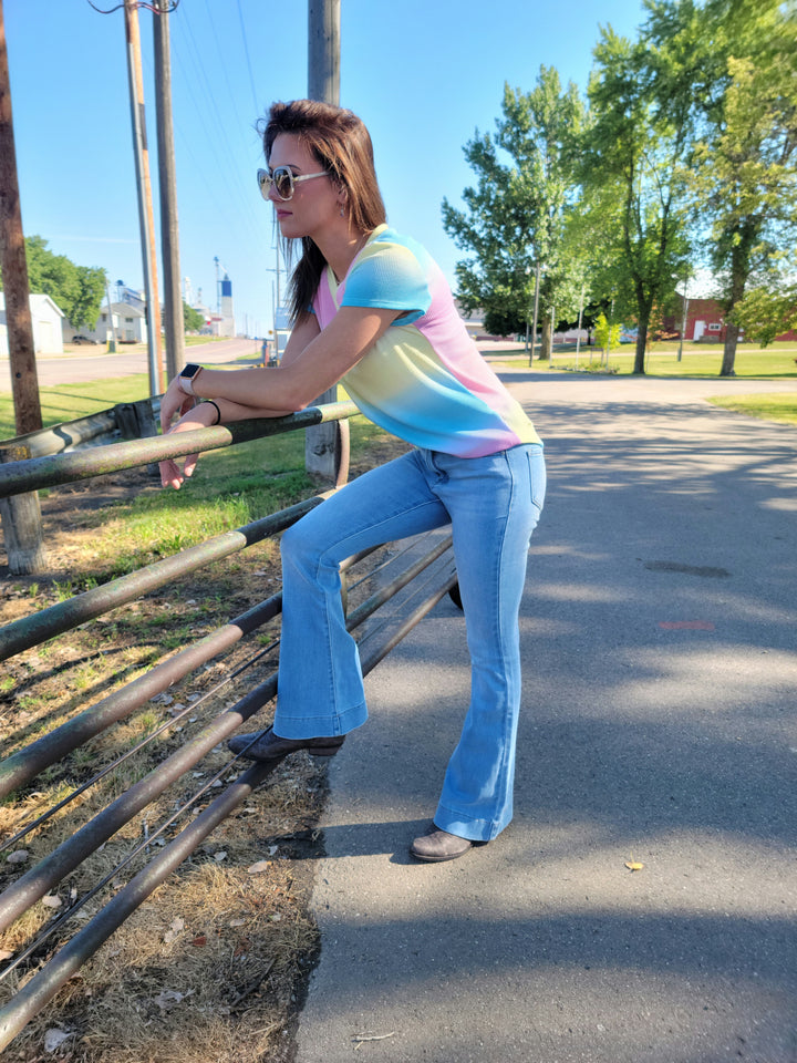 A woman in sunglasses and a pastel shirt wears KanCan’s Roxanne High Rise Flare stretch denim jeans, leaning on a metal gate outdoors with one foot propped up. Trees and a road can be seen in the sunny background.