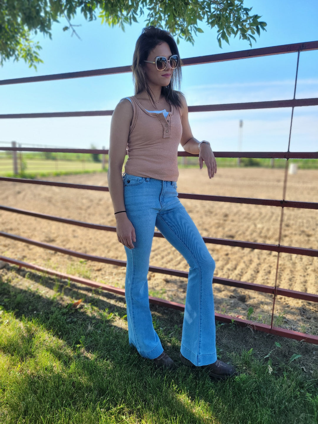 A woman in sunglasses, a sleeveless tan top, and KanCan’s Roxanne High Rise Flare jeans stands by a red metal fence on a sunny day, with a sandy riding arena and green grass behind her.