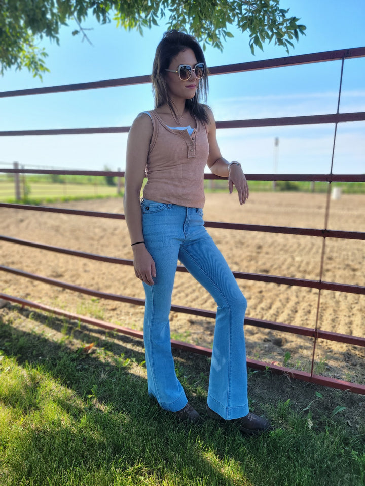 A woman in sunglasses, a sleeveless tan top, and KanCan’s Roxanne High Rise Flare jeans stands by a red metal fence on a sunny day, with a sandy riding arena and green grass behind her.