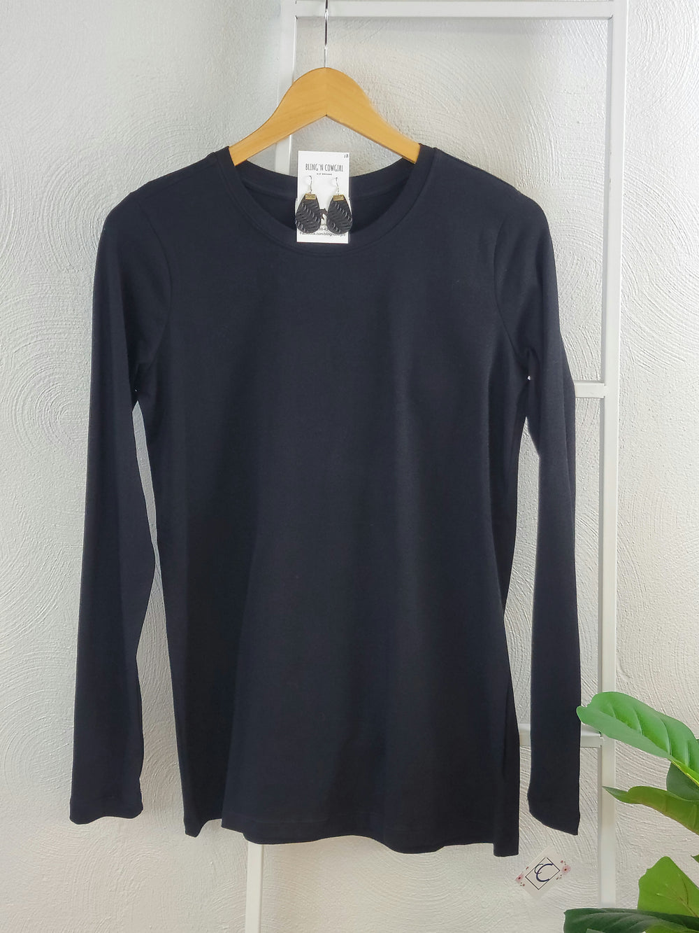 The Zenana Basic Long Sleeve Crew, a plain black crew neck shirt, hangs on a wooden hanger against a textured white wall with a small plant in the corner—perfect as a versatile layering piece.