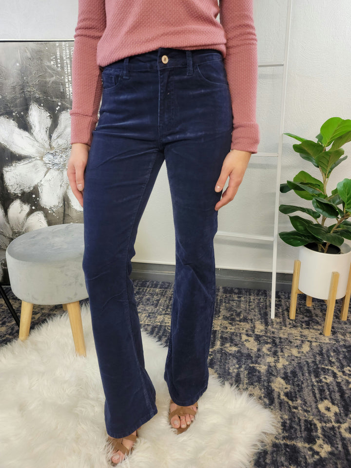 Wearing KanCan’s Genesis Corduroy Flares in high rise navy blue, a person with a textured pink long-sleeve top stands on a white rug. Their face is unseen; there’s a plant, modern stool, and flower painting in the background.