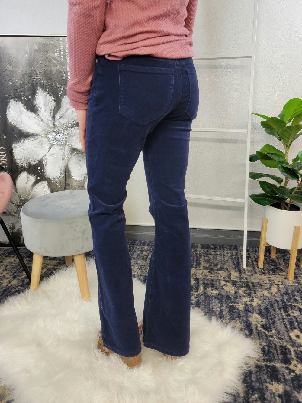 A person wearing KanCan's Genesis Corduroy Flares in navy blue and a textured pink top stands on a white furry rug, with a plant, small stool, decorative painting, and shelving unit in the background.