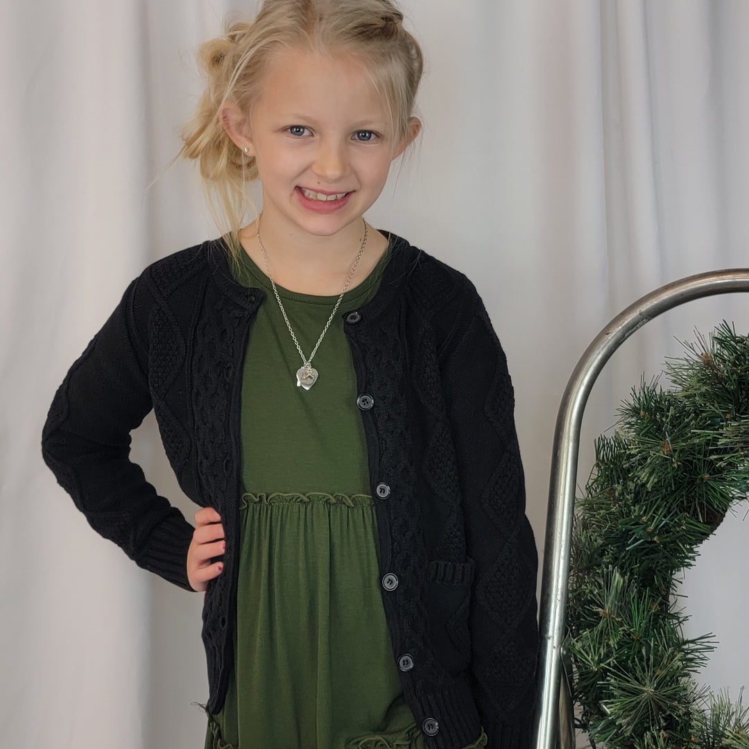 A blonde girl in a green dress and the Tele for Kids Shelby Cardigan smiles in front of a white backdrop beside a metal cart with greenery.