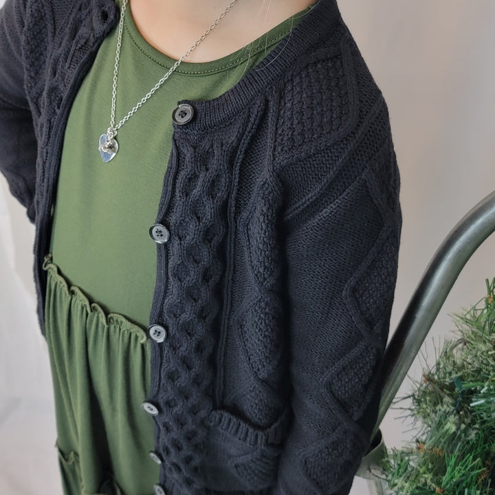 A child wearing the Tele for Kids Shelby Cardigan, a cozy black layering piece, over a green dress and silver heart necklace stands beside a decorated artificial Christmas tree.