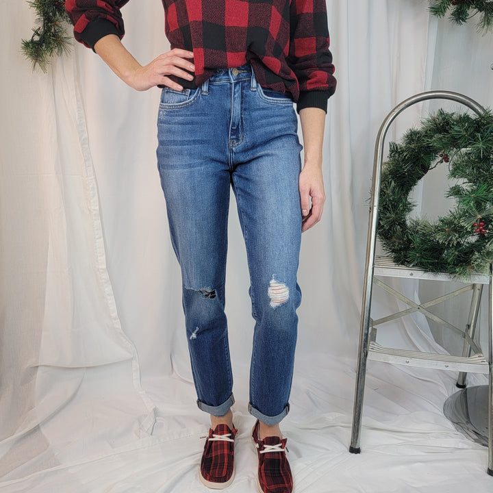A person wearing the Flying Monkey "Moon Legend" Mom Jean, a red and black plaid sweater, and plaid slippers stands indoors near a metal stepladder adorned with a pine wreath; white fabric drapes in the background.