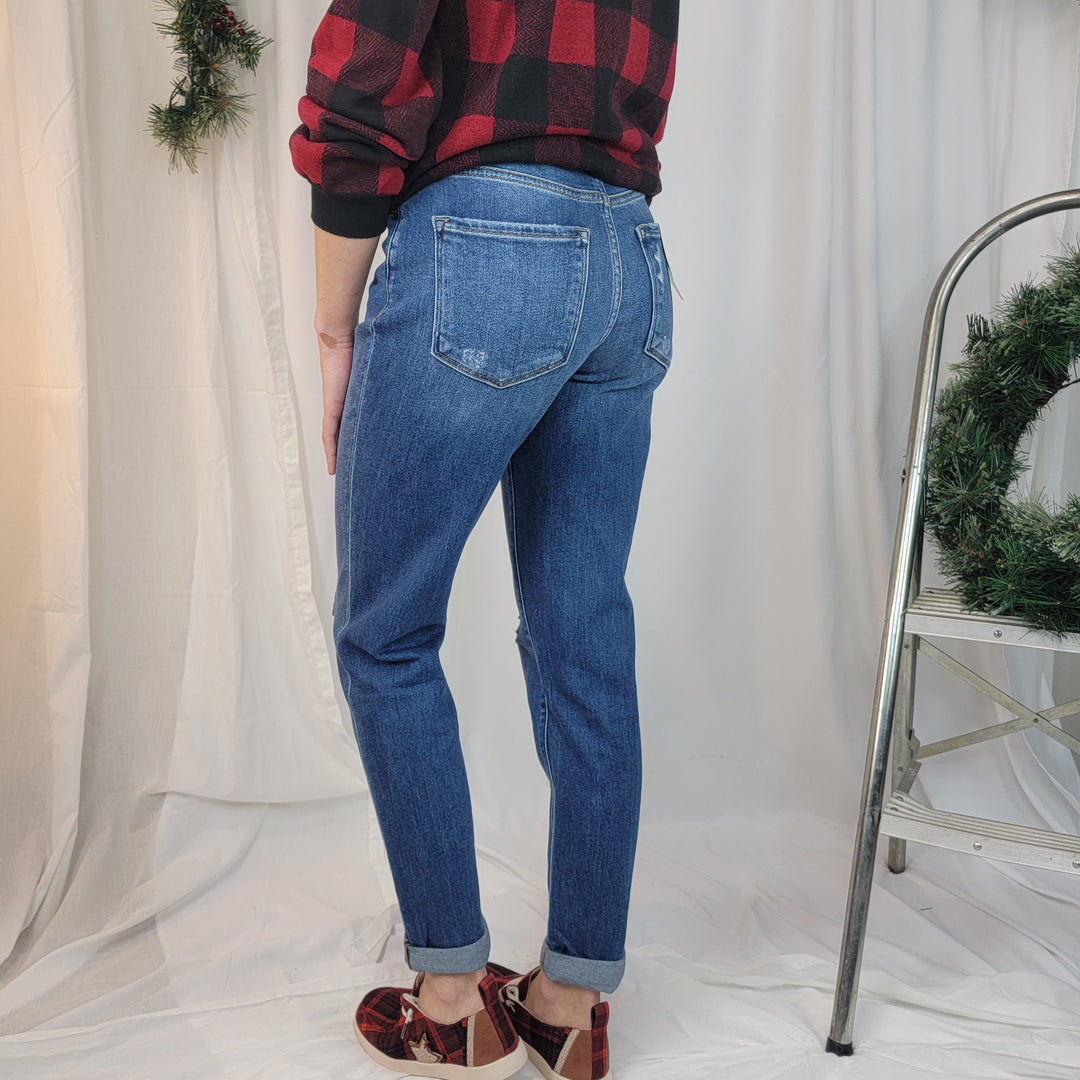 A person wearing Flying Monkey "Moon Legend" Mom Jeans, a red and black plaid shirt, and plaid slippers stands near a metal ladder with a wreath. White curtains and holiday greenery are in the background.