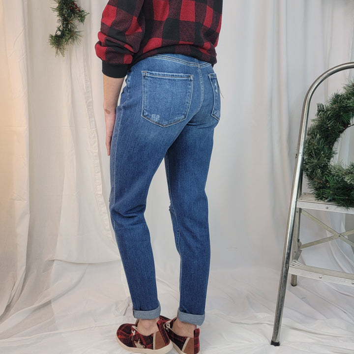 A person wearing Flying Monkey "Moon Legend" Mom Jeans, a red and black plaid shirt, and plaid slippers stands near a metal ladder with a wreath. White curtains and holiday greenery are in the background.