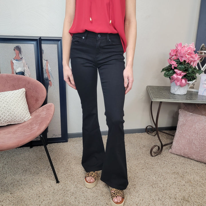 A woman in a sleeveless red top, KanCan Kimmy Black Flares, and leopard print platform sandals stands indoors by a pink chair, framed artwork, and a table with pink flowers.