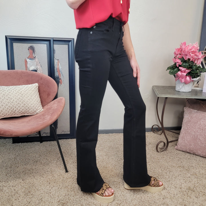 A woman wearing high-waisted KanCan Kimmy Black Flares, a red top, and leopard print sandals stands indoors near a pink chair, a side table with a vase of pink flowers, and framed art on the floor.