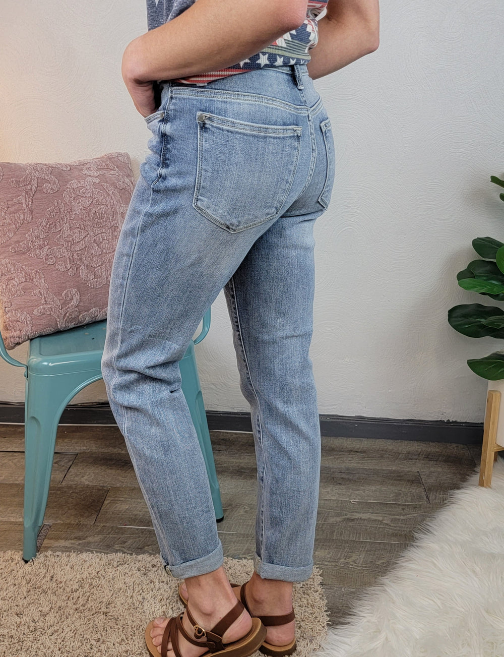 A person wearing Poppy Boyfriend Jeans by Judy Blue and brown sandals stands beside a green chair and pink pillow, with a leafy plant nearby on a wood floor with a white rug.