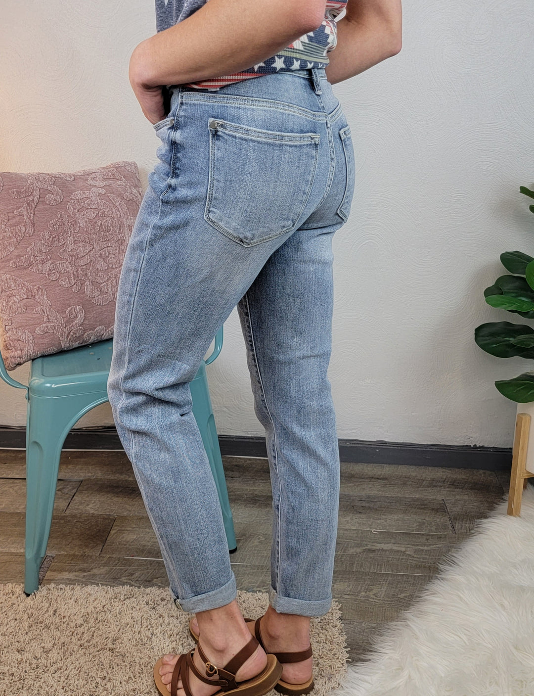 A person wearing Poppy Boyfriend Jeans by Judy Blue and brown sandals stands beside a green chair and pink pillow, with a leafy plant nearby on a wood floor with a white rug.