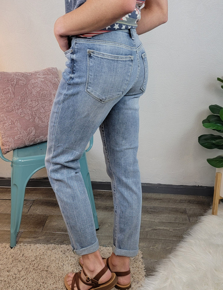 A person wearing Poppy Boyfriend Jeans by Judy Blue and brown sandals stands beside a green chair and pink pillow, with a leafy plant nearby on a wood floor with a white rug.