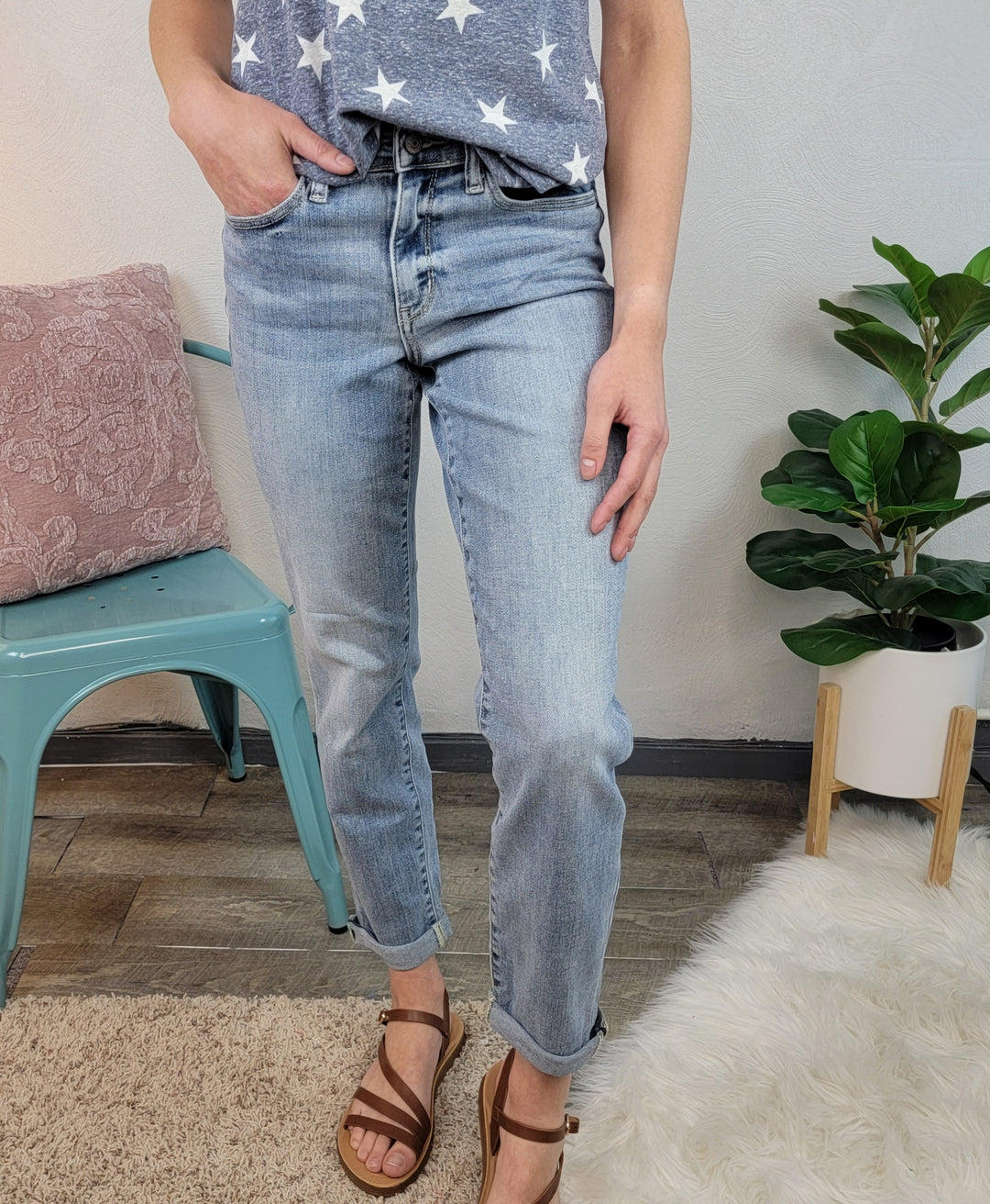Someone wearing the Poppy Boyfriend Jeans By Judy Blue, a navy star-patterned shirt, and brown sandals stands indoors near a green chair, potted plant, and white furry rug; their face is not visible.