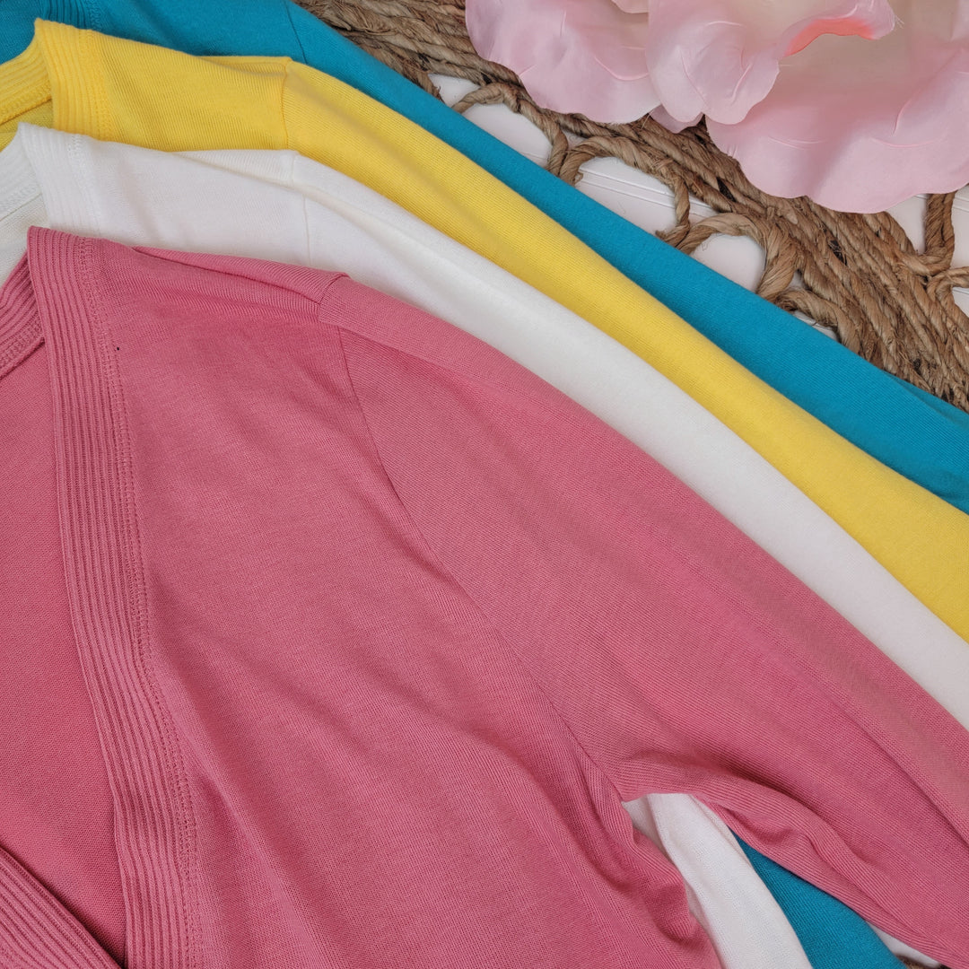 Four Long Sleeve Snap Front Cardi sweaters by Zenana in pink, white, yellow, and teal are neatly stacked on a woven mat with artificial pink flowers nearby. Available in extended sizes.