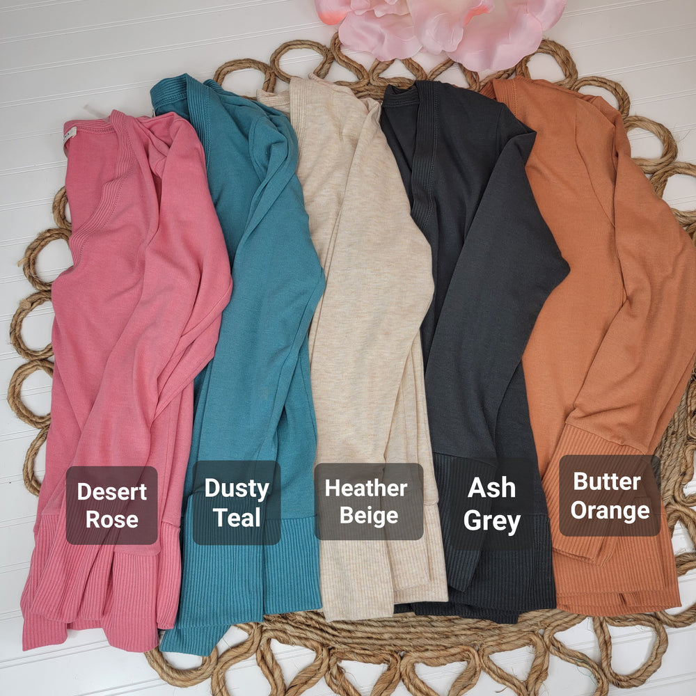 Five Zanana Long Sleeve Snap Front Cardis in Desert Rose, Dusty Teal, Heather Beige, Ash Grey, and Butter Orange are neatly folded on a rope mat—perfect cozy layers to add to your wardrobe.