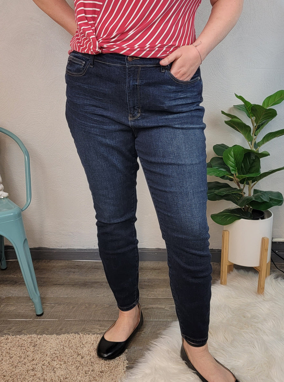A person in a red and white striped top, Mandy Skinny Jeans by Judy Blue, and black flats stands indoors with one hand in a pocket. A green plant and a blue chair are in the background.